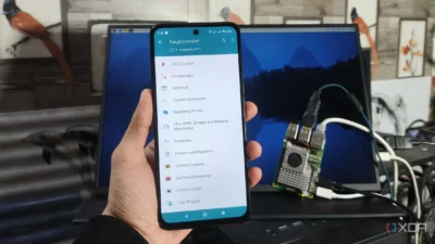 The image shows a hand holding a smartphone displaying the RaspController app, which appears to be used for managing a Raspberry Pi. In the background, there is a computer monitor showing a dark blue screen with what seems to be a landscape image, and a framed picture of a bird on the wall. The foreground mainly consists of the smartphone and a part of the hand holding it. The setting appears to be an office or workspace. The smartphone is displaying the app's interface with various options relating to the Raspberry Pi's functions, including GPIO control, file management, SSH shell access, and camera settings. A small detail shows that a Raspberry Pi 5 is connected and being controlled.