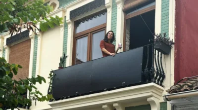 The photograph shows a woman standing on a balcony, holding a cup. The balcony is attached to a building with old-style architecture, featuring decorative moulding above the windows and a mix of light beige and dark teal colours on the façade. In the foreground is a black solar panel that looks almost like a privacy screen that extends the length of the balcony. Plants are visible in pots both on the balcony and in the background growing along the building's exterior wall.