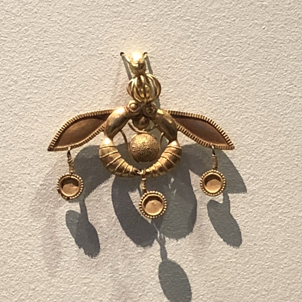 Gold jewel with two honeybees touchions heads and tails to form a circle, with a ball of pollen (or a drop of honey) in the centre.