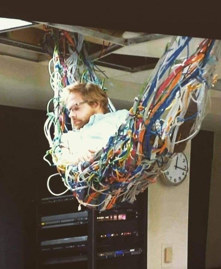 a man with glasses lying in a mess of wires like it's a hammock, as it hangs from the ceiling in an office building