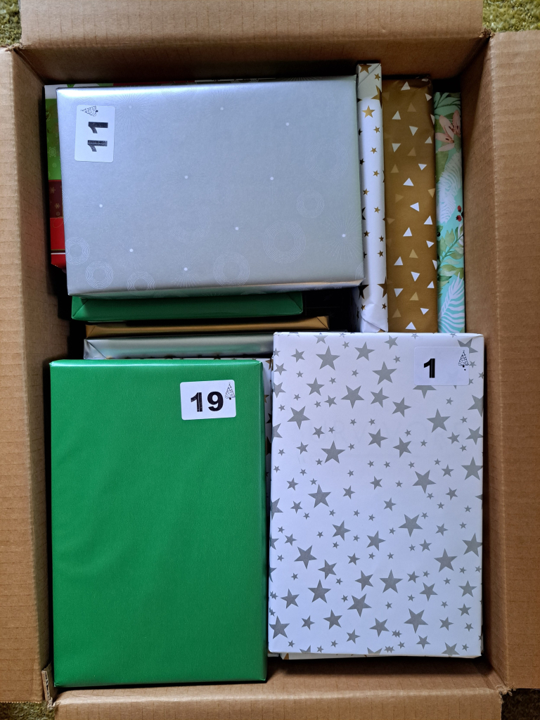 A cardboard box filled with wrapped books. Of the books that can be seen, the wrapping paper is either green, white with gold stars, silver, gold with white triangles. Each book has a white sticker with a number denoting the date it is to be opened.