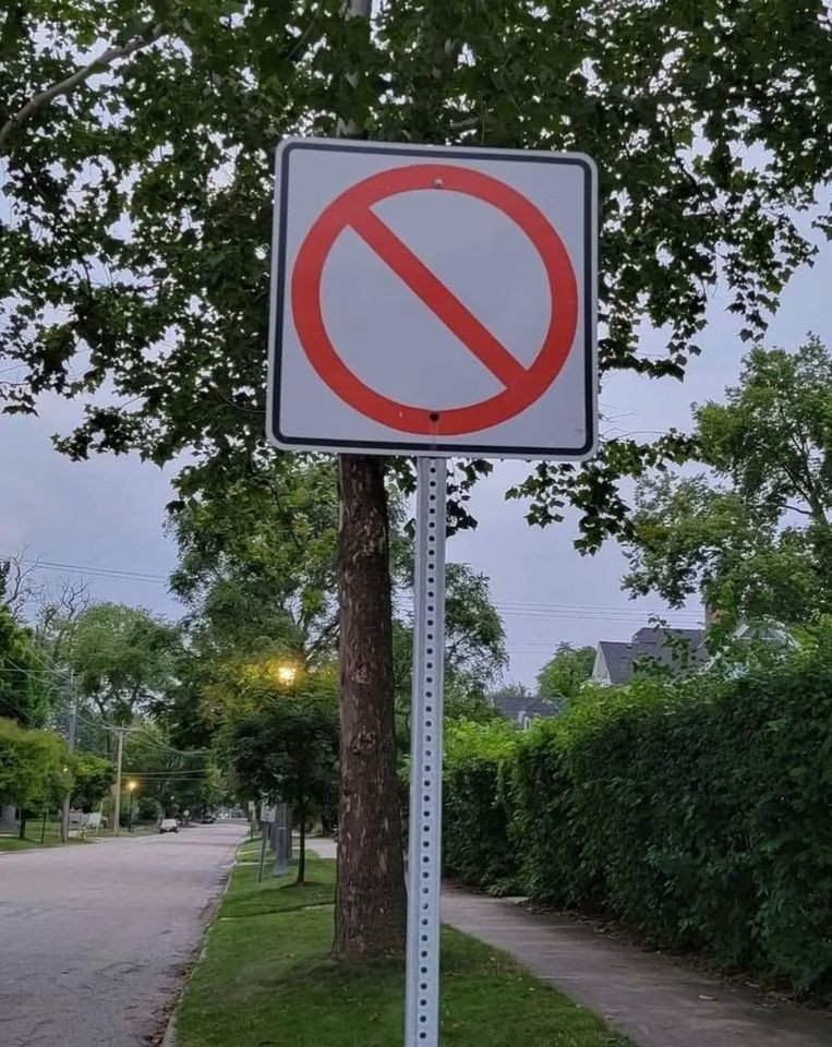 suburban street with trees and a hedge, and a prominent sign that has the red circle with slash through it, but on a white background