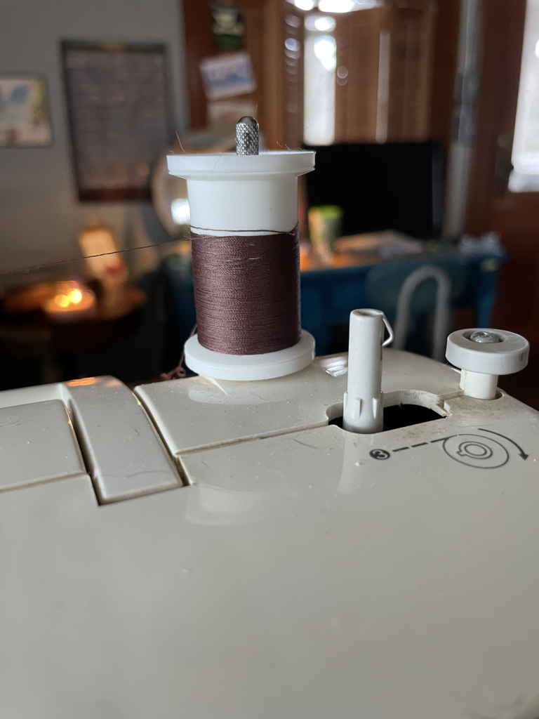 A spool of brown thread on a sewing machine. The spool is nearing the end with about 1/3 of white spool showing at the top. 