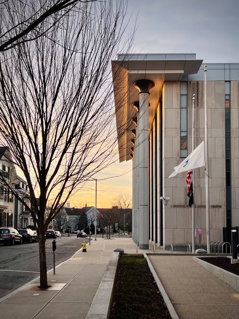 Looking down a sidewalk, the facade of a modern courthouse building is illuminated by late afternoon sunlight. 