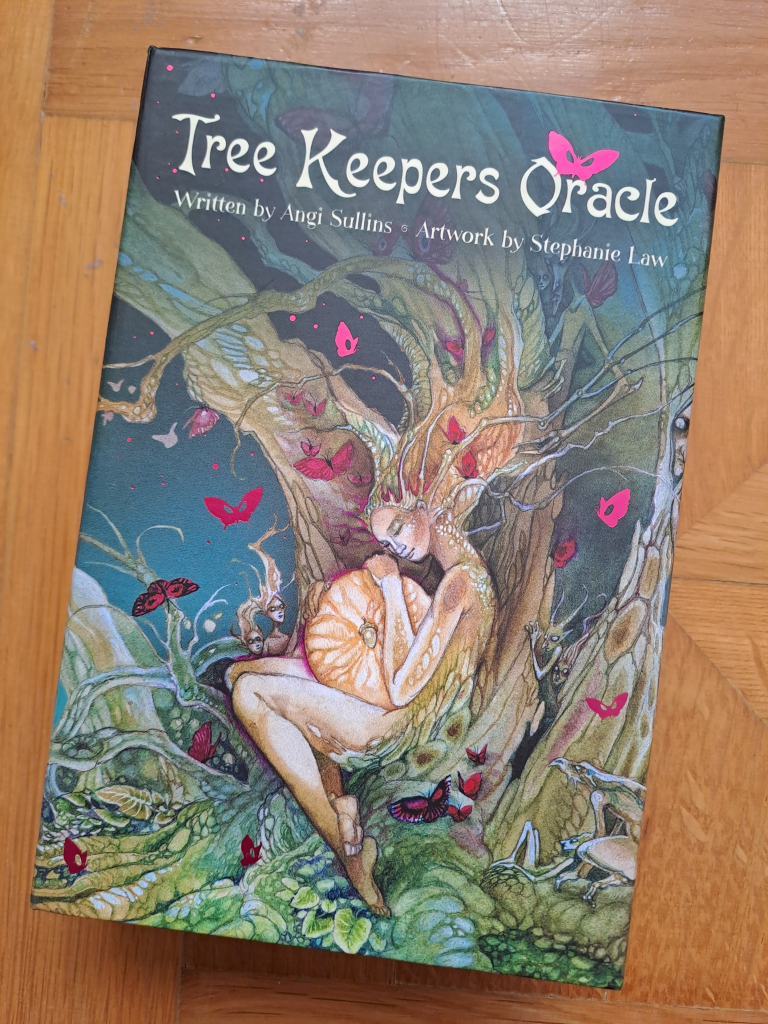 Box of the Tree Keepers Oracle deck, written by Angi Sullins and illustrated by Stephanie Law. It shows a beautifully detailed, colorful watercolor image with a woman curled around a glowing orb at the base of the tree, peacefully sleeping, half merged with the tree bark, hair growing into branches. There are bright pink butterflies flitting around. 