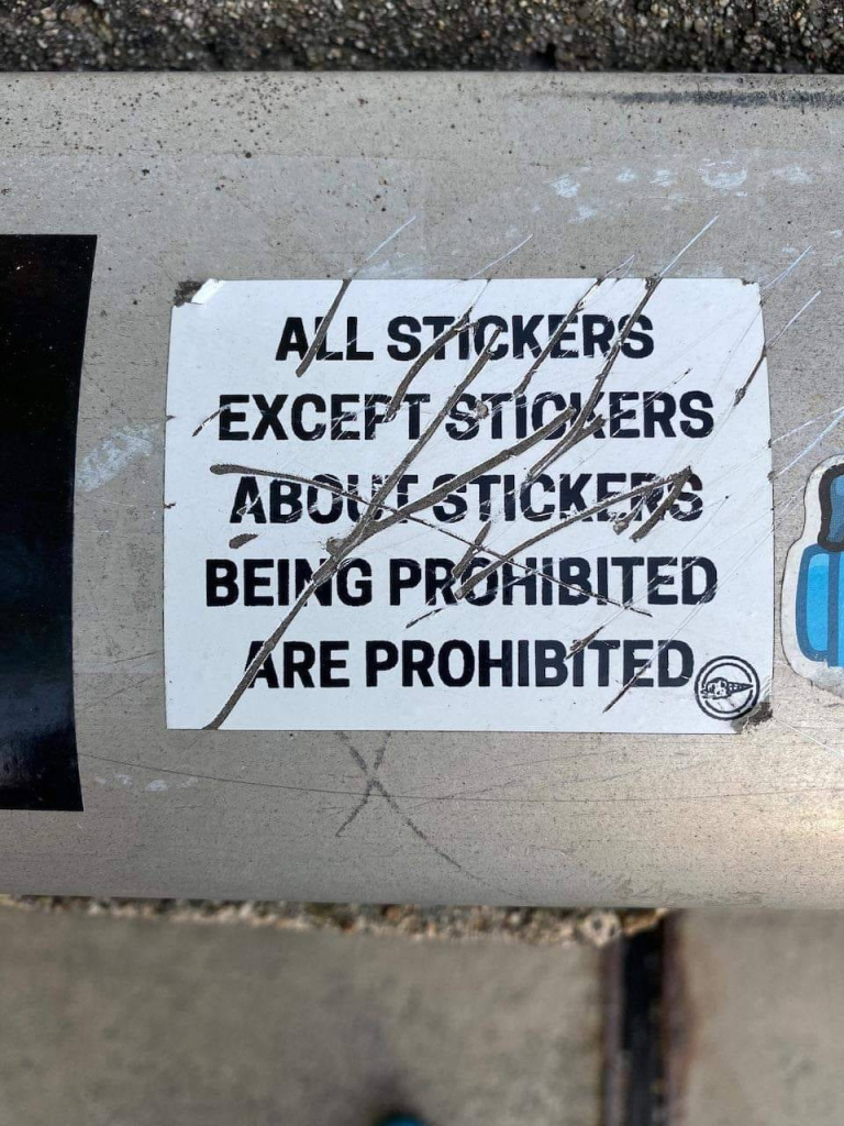 sticker on a curb with the text scratched up: All stickers except stickers about stickers being prohibited are prohibited