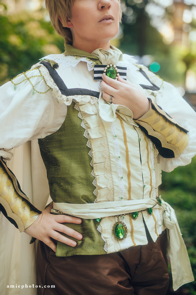 This is a close up of my toso in the costume. I am holding the giant acrylic gemstone that is at my neck. There are more around my waist. There’s a lot going on. The vest is green silk with a panel down the front. The vest is trimmed in lace and rhinestones. 
Watermark reads amiephotos.com