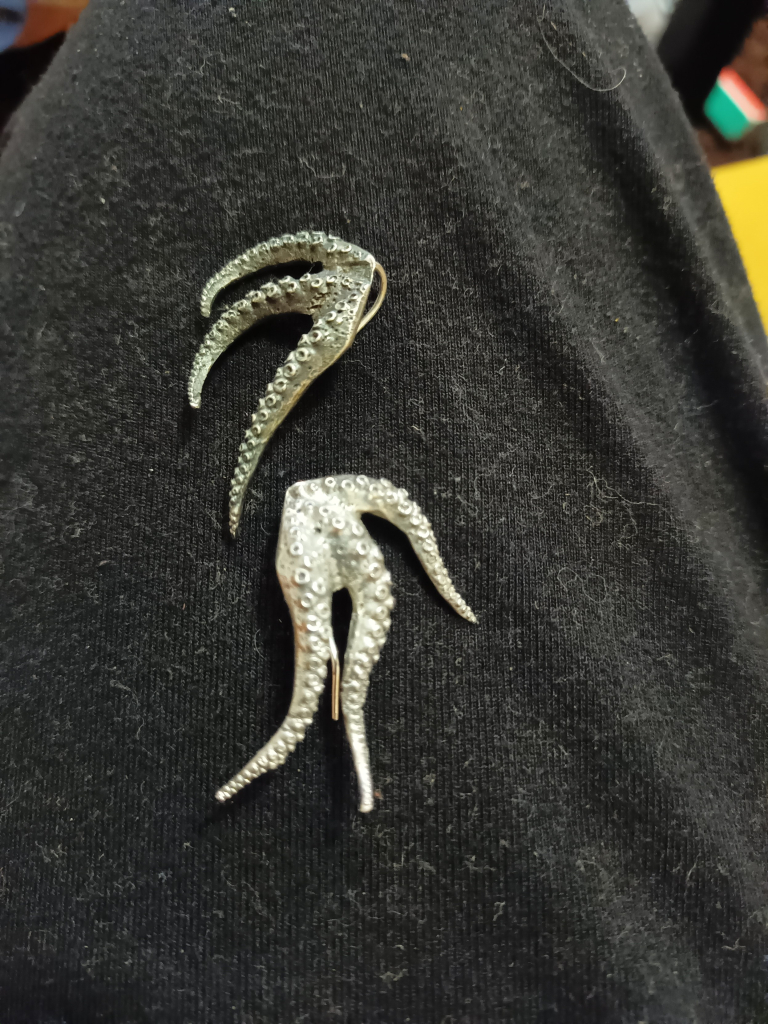 Two silver earrings, each made of three tentacles with many tiny suckers. One earrings is polished and the other is noticeably darker.