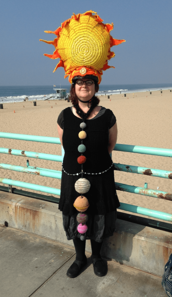 A woman (me) standing on a pier at the beach, wearing a crocheted solar system costume, including a large yellow sun-shaped headpiece and a black tunic, and a line of crocheted balls representing the planets, secured at the waist with a glittery beaded belt.