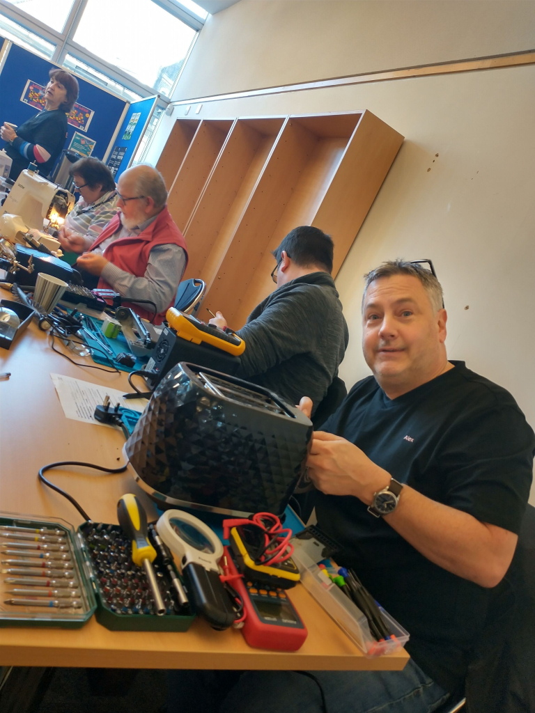 Alex, a man with short grey hair and glasses on top of his head, looks at the camera while working on a toaster. Around him on the table are lots of bits of fixing equipment. Behind him are several other volunteers all working on different things at other tables.