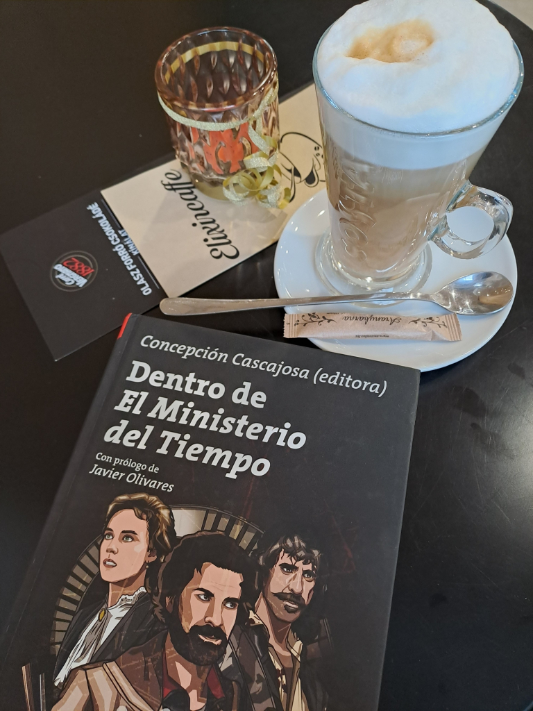 A coffee table with a cup of latte, a tea candle, and a copy of the book "Dentro de el Ministerio del Tiempo" edited by Concepción Cascajosa. Featuring comic book style images of the three main characters in the series.