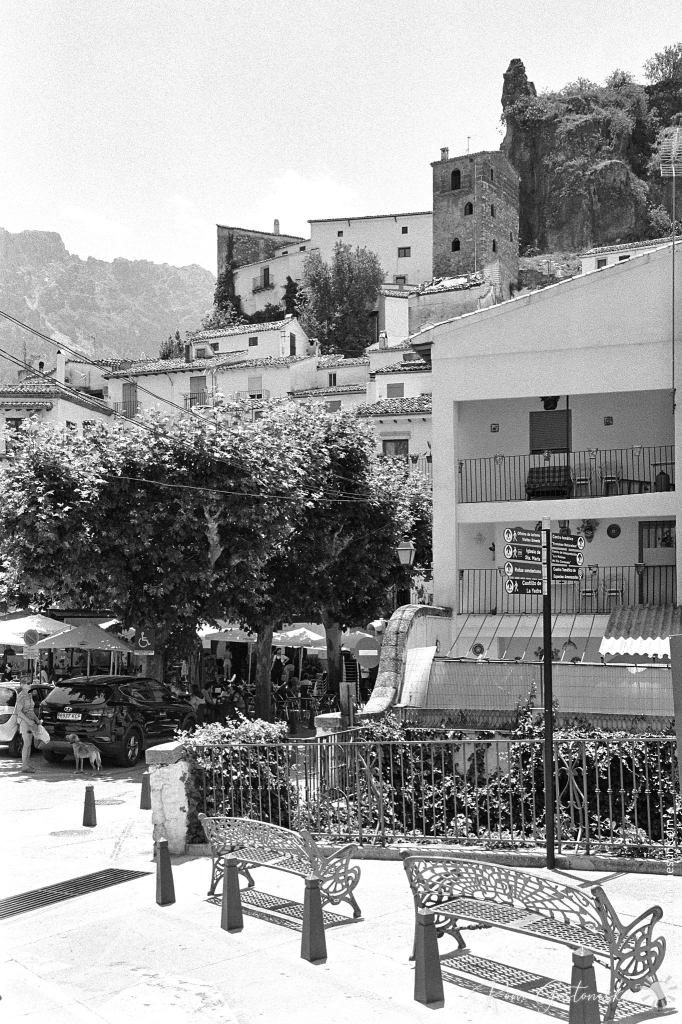 A black-and-white photo of two iron benches in the foreground. To the left is the oldest of the three squares in Cazorla. Beyond that, houses rise vertically on an almost vertical cliff, seemingly defying gravity.