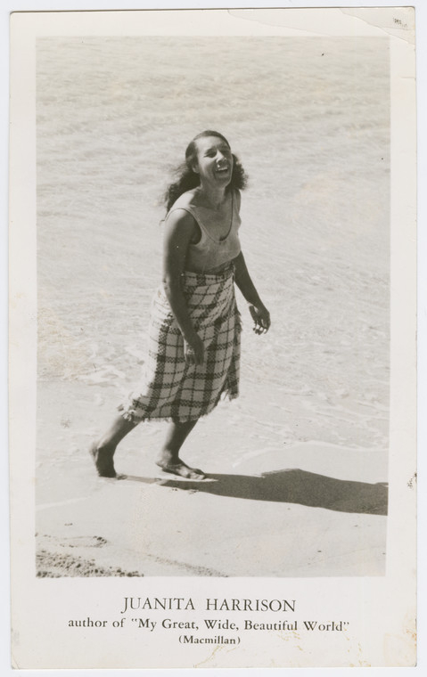 A black and white photo of Juanita walking barefoor on the beach. She is wearing a plaid wrap skirt and a sleeveless blouse, with her hair loose and swept back, and her face is lit up with a bright radiant smile. Under the photo there is an inscription: "Juanita Harrison. Author of My great, wide, beautiful world (Macmillan)"