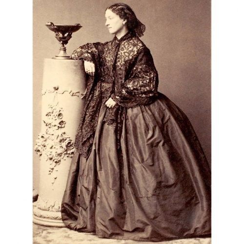 Black and white photograph of Jeanne Villepreux-Power dressed in a wide skirt and a lace shawl wrapped over her shoulders. She is leaning on a decorative column, looking at an elaborate cup. She is young and elegant, with her hair pulled back in a net.