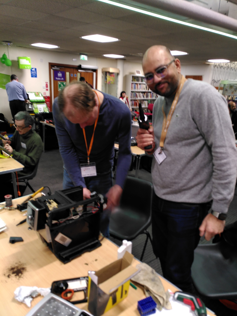 Two volunteer repairers fixing a coffee machine. One is engrossed in what they are doing, the other is looking to camera and is giving a thumbs up.