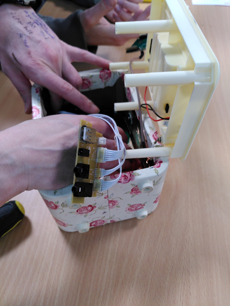 One of our volunteer repairers fixing a radio. The photo only shows a close-up of her hands as she is reaching inside it. One of her hands is covered in notes to self written in biro. The radio is in a kind of white and pink chintzy case.