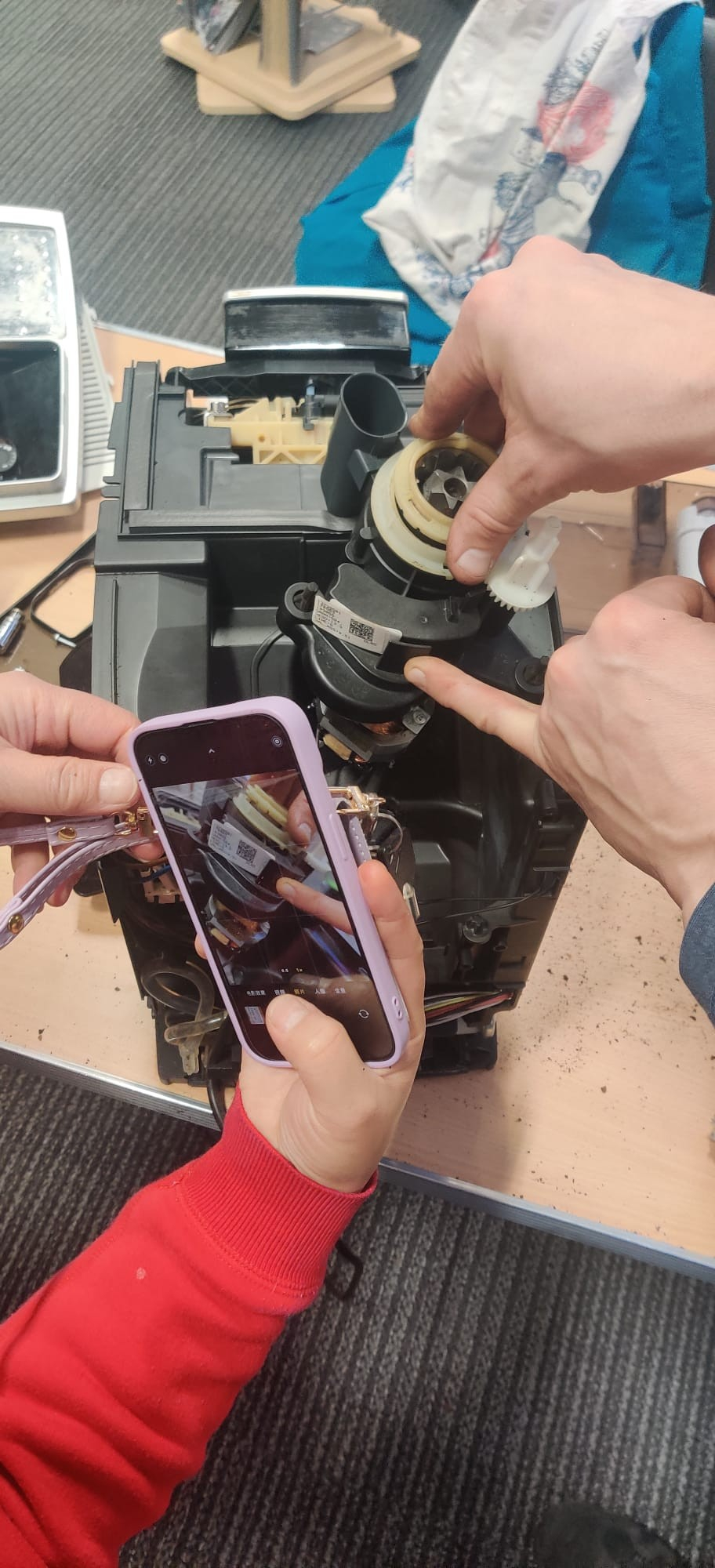 Close-up of repair to a coffee machine. The owner is holding a phone to take a picture of the inside while the volunteer repairers is pointing at a particular part.