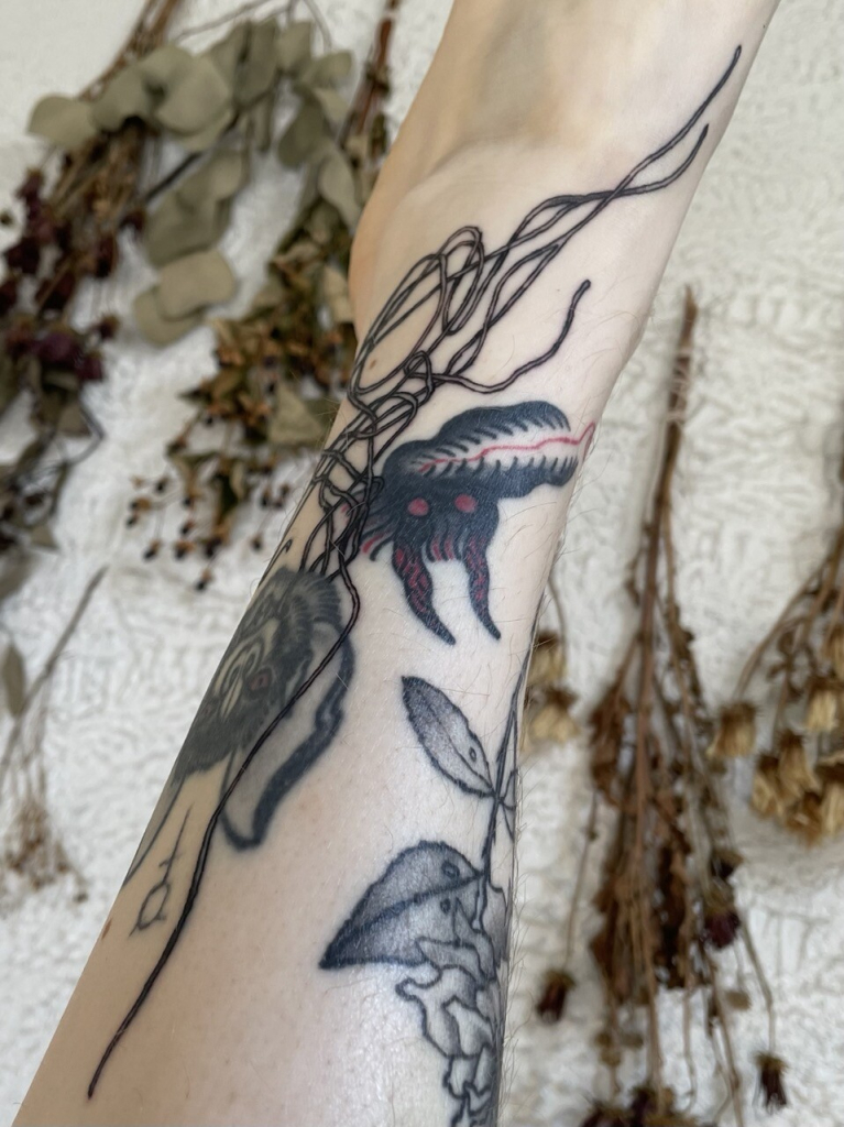 black and grey tattoo of abstract lines on an ankle, entertwined with older tattoos. in the background are bouquets of dried flowers hanging on a stuccoed wall