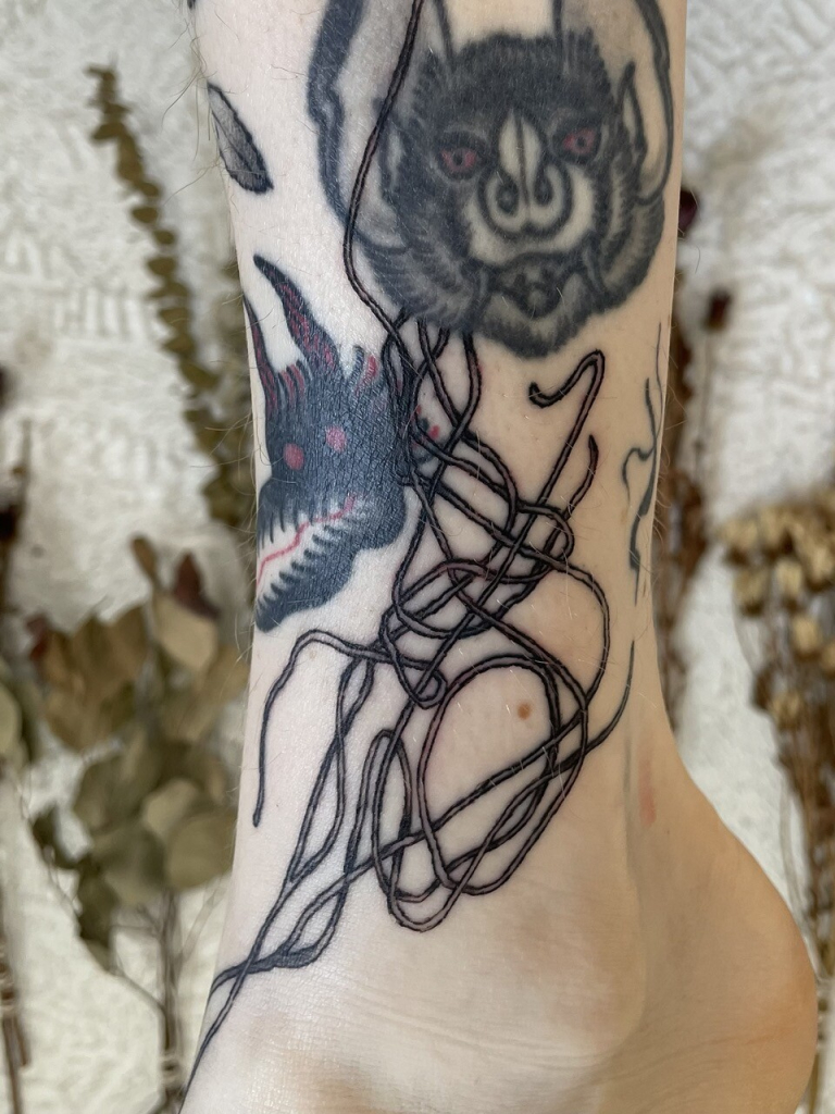 black and grey tattoo of abstract lines on an ankle, entertwined with older tattoos. in the background are bouquets of dried flowers hanging on a stuccoed wall