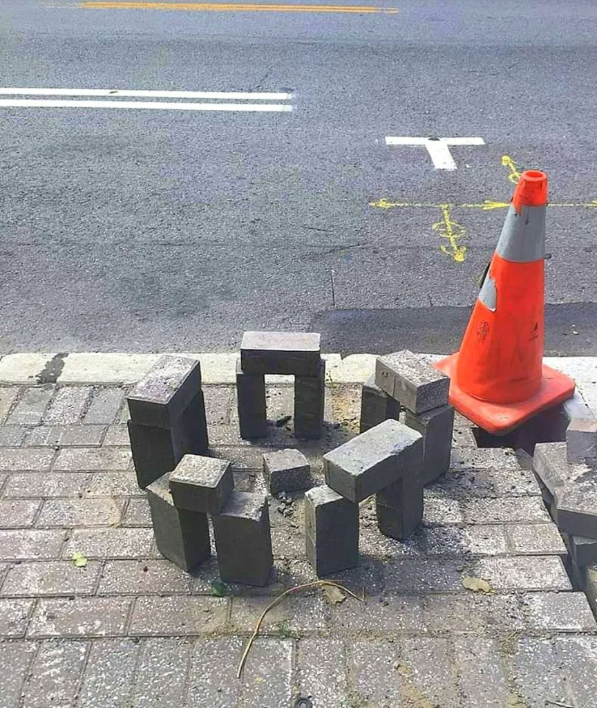 Tiny replica of stonehenge, built out of paving bricks on the sidewalk. There is an orange traffic cone looming next to it.