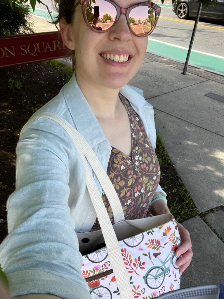 A selfie of a Caucasian woman smiling at the camera. She is wearing reflective rose gold sunglasses. Her dress has a v-neck and is a brown jersey fabric printed with raspberry leaves and brambles, with a pale blue fitted linen overshirt.  Over her shoulder is a cream-colored tote bag with cotton straps, printed with autumn leaves and teal bicycles. 