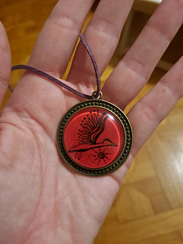 A round, bronze colored pendant with an image under a glass lens in the middle. The image is a stylized bird in flight, on a red background. The pendant is in the palm of my hand, 3cm in diameter.