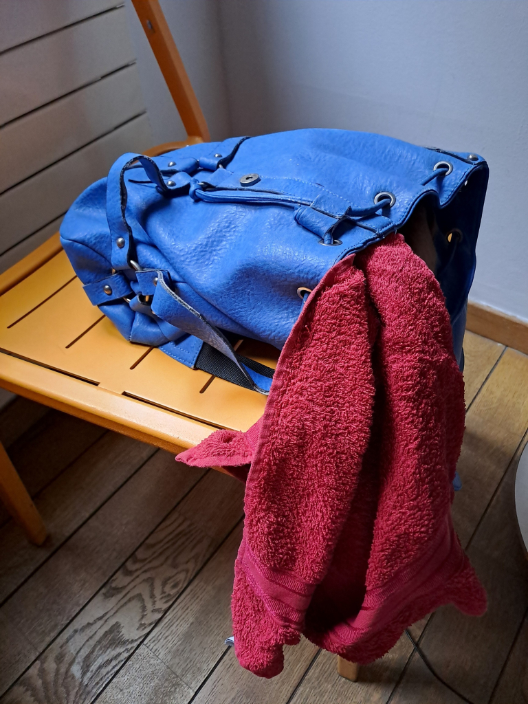 A small blue backpack placed on an orange metal chair. A red flannel towel is hanging out of the open bag.