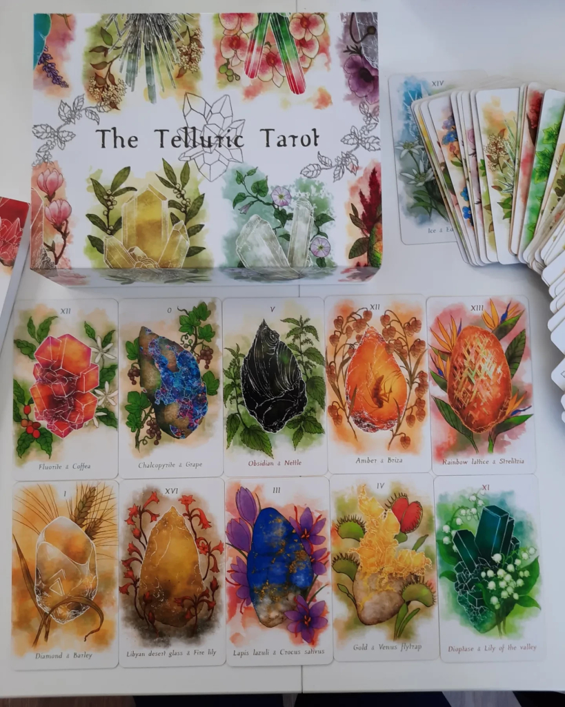 The box of Telluric Tarot with a few cards spread out. It is a vividly colored watercolor style deck. Each card has a mineral and a plant on it, which combine their symbolism to represent the card. E.g. Obsidian and Nettle.