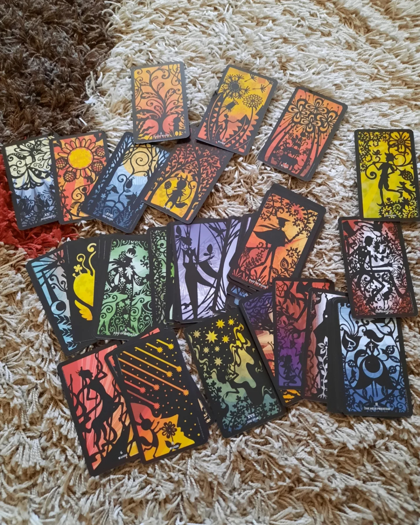 Silhouettes Tarot cards scattered on the carpet. They are black, paper shadow theater style silhouette designs of whimsical scenes, with brightly colored backgrounds.