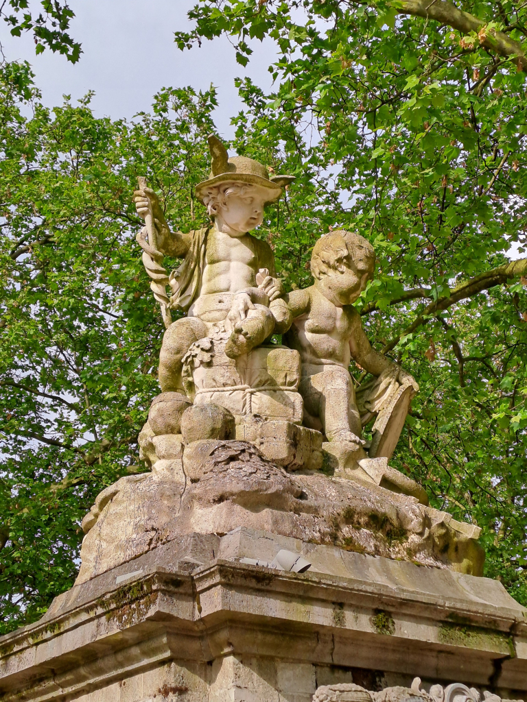 A stone statue in a park, featuring two small children sitting next to each other. One of them is wearing Hermes' hat and holding a serpent staff, and has very well defined abs and pecs. 