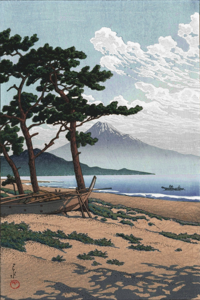 A view looking out over a sandy beach toward a distant Mount Fuji, on a bright afternoon. In the foreground, irregular patches of grass cling to the sandy beach; three tall, wiry pine trees stand at a permanent angle, sculpted by the wind coming in off the water. A small boat has been dragged up the beach and tucked in beneath the pine trees; further on, out on the calm blue water, another similar boat carrying a pair of passengers approaches the shore. Low green hills line the far side of Suruga Bay, while futher on, snow-capped Mount Fuji stands tall, its base lost in colourless mist, framed by bright blue sky and ragged clouds making a sweeping arc out over the ocean