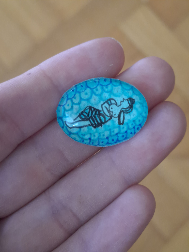 An oval glass pendant on my palm, 2cm wide. It features the black line drawing of a stylized Minoan figure, a woman in a flounced skirt and bare breasts reclining against a blue and green background of stylized waves.