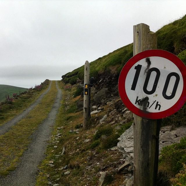 a rough track on the side of a hill leading to a steep decline, in ireland, with a speed limit sign in the foreground that says 100 km/h