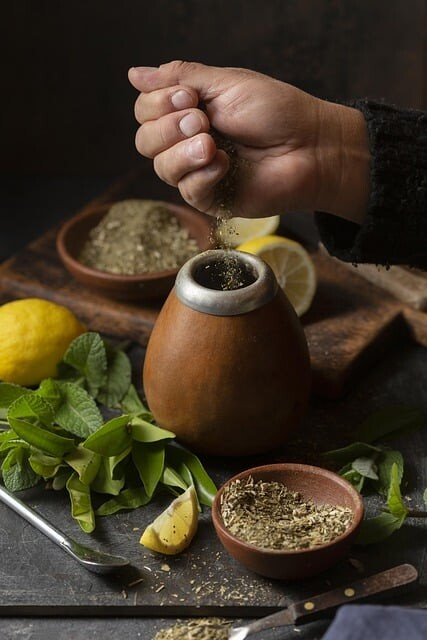 A photo of a hand pouring ground spice or tea into a little wooden vessel. Lemons and different kinds of leaves are arranged on a stone cutting board around it with a bowl or mortar containing more of the spice or tea.