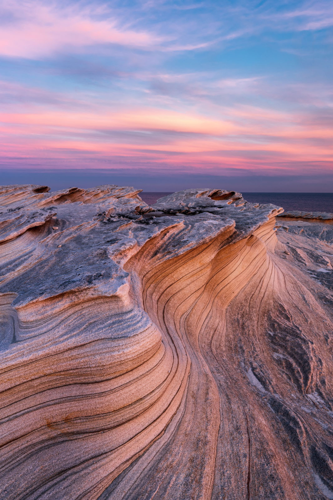 a vertical sandstone landscape with sandstone looking like a wave ahd making two curves. The photo is showing sunset time so both the sky and the rocks are in pink tones.