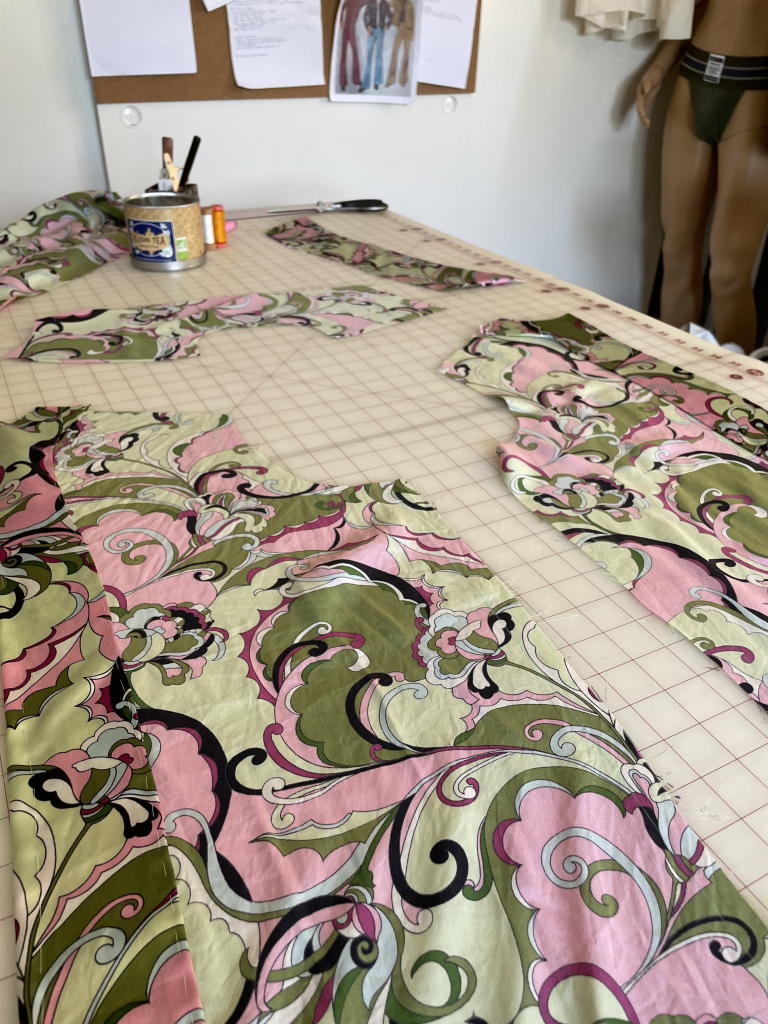 Front, yoke, and collar pieces of a shirt lay flat on a gridded cutting table. The fabric is a vintage 1960s floral print silk in greens and pinks. 