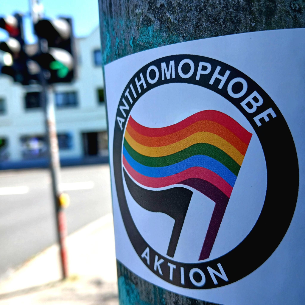 Das Bild zeigt einen Aufkleber, der an einem Pfosten befestigt ist. Der Aufkleber zeigt die Aufschrift "ANTIHOMOPHOBE AKTION" in einem schwarzen Ring, der den äußeren Rand bildet. Im Zentrum des Aufklebers befindet sich eine Fahne mit den Farben des Regenbogens, die ein Symbol der LGBTQ+ Gemeinschaft darstellt. Der Hintergrund zeigt unscharf eine Straße mit einer Ampel und Gebäuden im Freien, was auf eine städtische Umgebung hindeutet. Die Farben auf dem Aufkleber sind lebendig, und das Bild wurde bei Tageslicht aufgenommen.