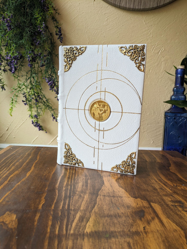 A white leather journal sitting upright in a desk. The cover has a good coin in the center, gold circles, and corner protectors.