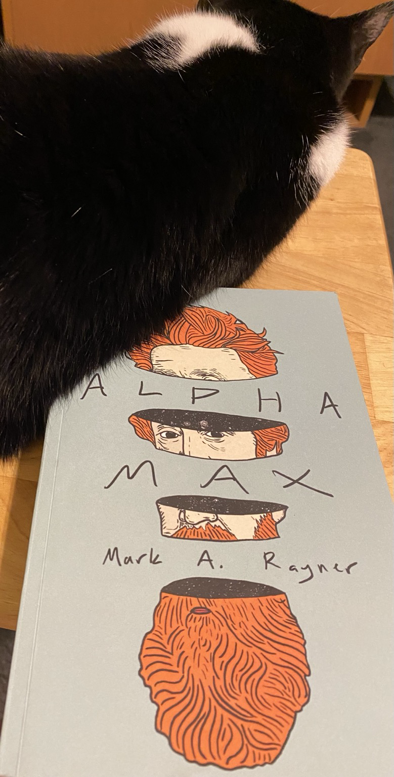 A copy of the book Alpha Max, being pushed off a tray table by a cat who has called dibs on the space. 