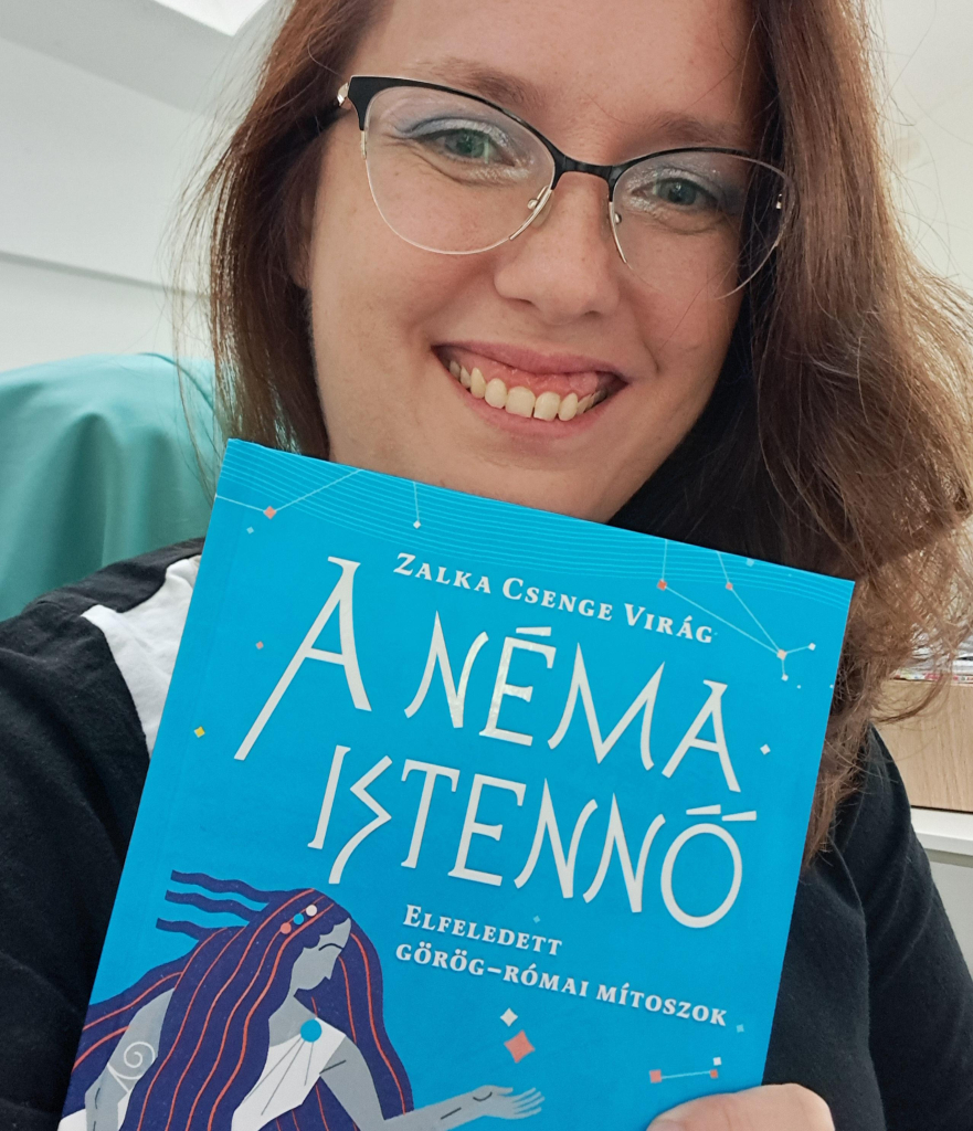 Photo of me (white woman, 30s, long red hair, glasses, blue eyes, sparkly makeup, toothy smile), holding a book with a blue cover, decorated with constellations and the stylized image of a Greek goddess with long flowing hair. Title (in Hungarian) reads The Mute Goddess: Forgotten Greek and Roman Myths.