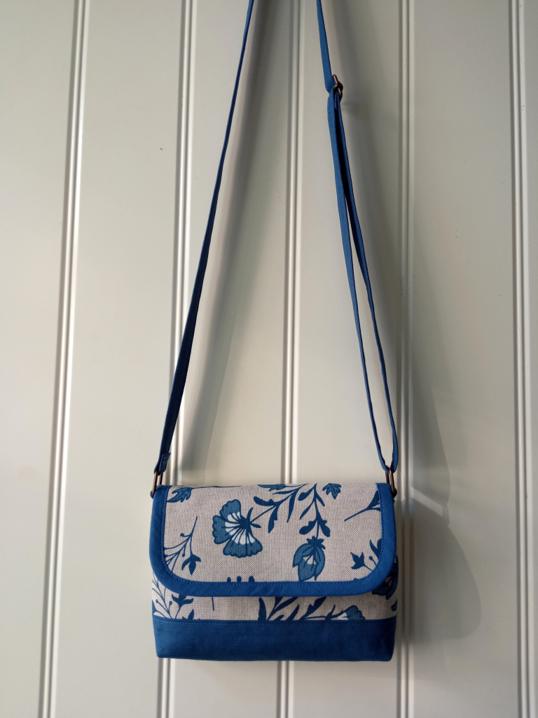 On an offwhite shiplap door a small handbag is hanging. It's beige with a floral pattern in navy, a lighter blue and white. The bottom, the strap and bias binding of the flap are blue.