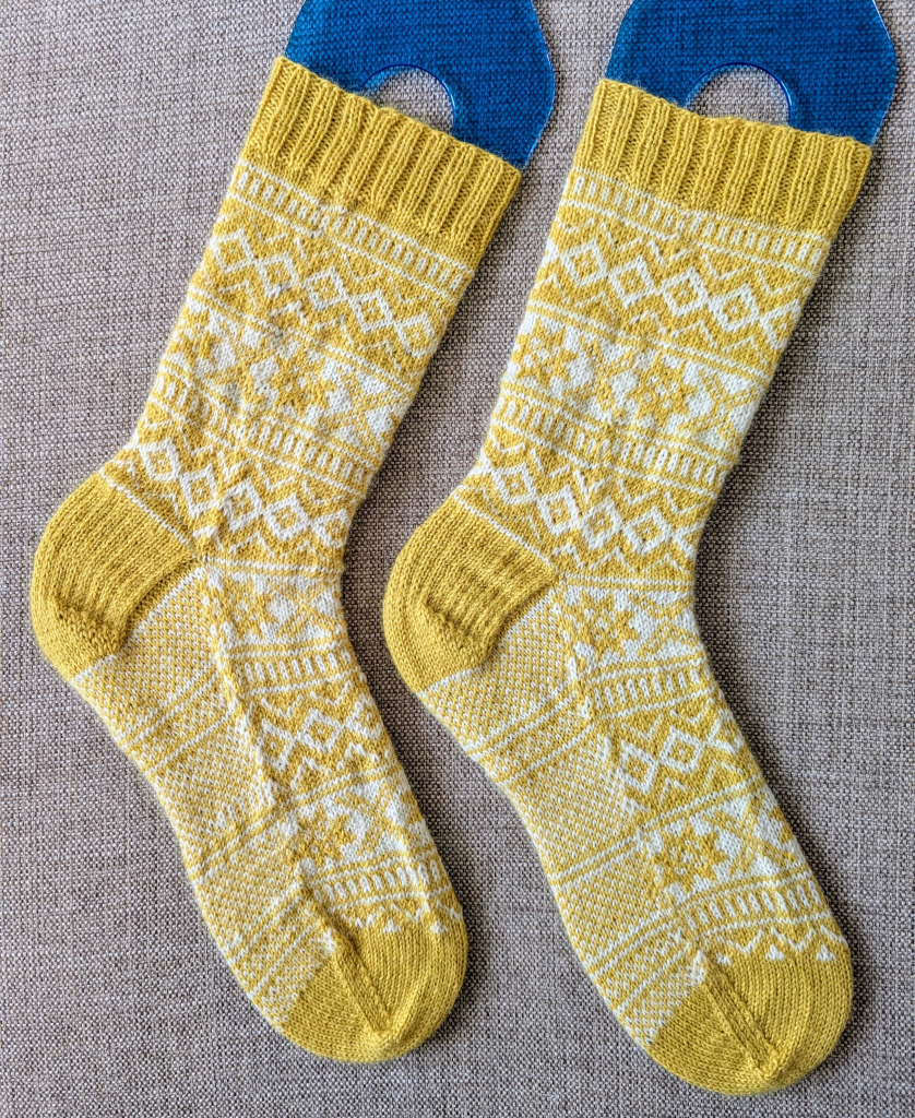 A side view of a pair of yellow and white knitted socks, with repeating bands of stranded colourwork. The motifs are a mixture of diamonds, X's, and snowflakes. The sole of the socks is a basic checkerboard pattern. 