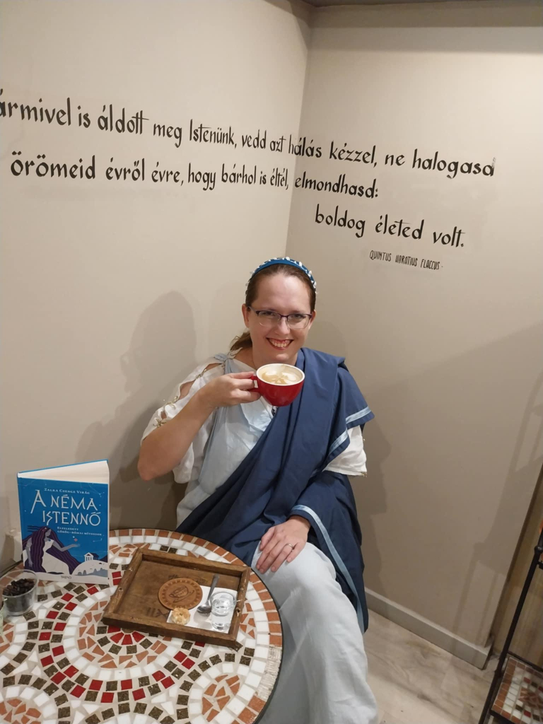 Me (woman in her thirties, red hair, glasses) sitting in the corner of a café, drinking a cup of latte. The coffee table has a mosaic theme, and there is Horatius quote on the wall behind me. My book, The Silent Goddess: Forgotten Greek and Roman myths, is on the coffee table. The cover is blue, with art featuring a goddess surrounded by stars. I am wearing a white and blue Roman outfit and a blue hairband.