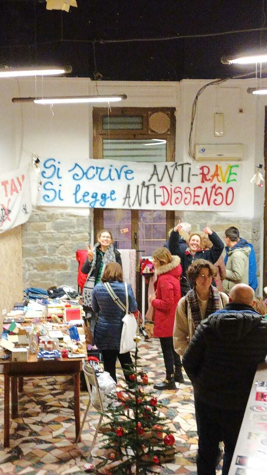 People in a well lit room. Items lie on tables. Political banners hang on the walls. A small Christmas tree is in the foreground. Cheerful!