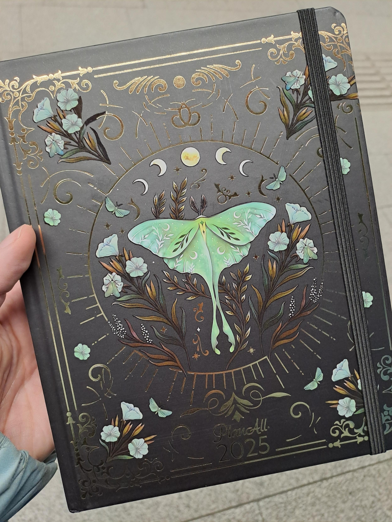 A hardcover planner with a black cover that features an intricate gold and green design. The center of the design is a green luna moth, surrounded by lunar symbols and flowers as well as gold motifs.