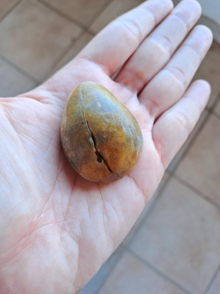 Photo of a pebble that fits in the center of my palm. It is egg shaped, smooth, with spots of orange, grey and brown. There is a line down the middle lengthwise where the pebble is split, but the two halves fit perfectly together.