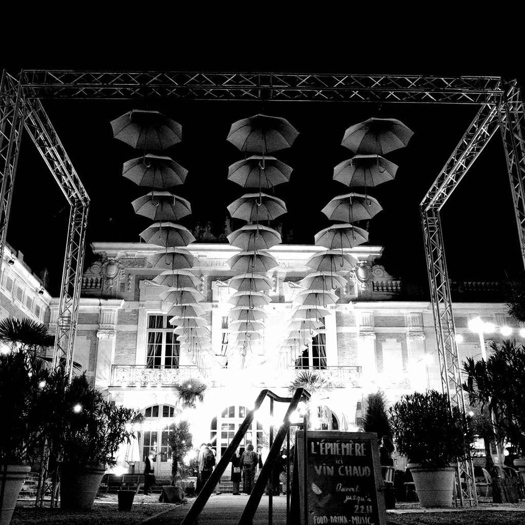 Photo de parapluies installés sur des câbles tirés entre le mur d'un bâtiment et des montants métalliques. Une lumière éclaire le théâtre et apparait vive dans le décor nocturne.