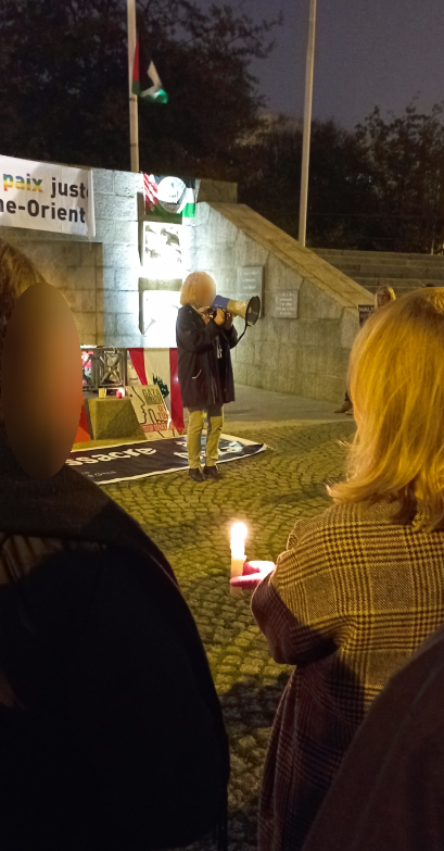 Une personne fait un discours dans un mégaphone dans une ambiance nocturne, devant le monument aux morts de Brest. À l'arrière plan, on voit des drapeaux palestiniens et libanais.