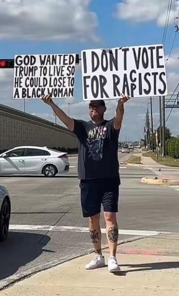 Man on the side of the road holding two signs: 

GOD WANTED TRUMP TO LIVE SO HE COULD LOSE TO A BLACK WOMAN

I DON'T VOTE FOR RAc/pISTS