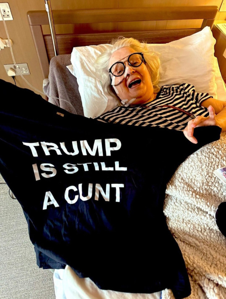 Comedian Janey Godley is lying in bed, intubated. She has an open-mouthed smile and is holding up a black T-shirt with bold, white which reads: "Trump is Still a Cunt".